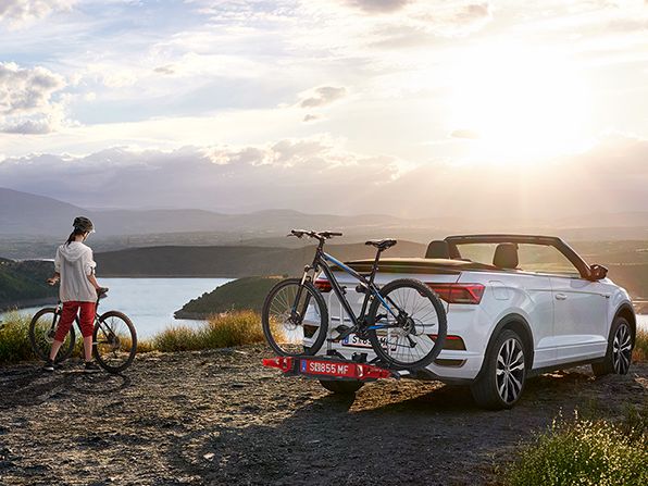 Ein VW T-Roc Cabrio in weiss mit Fahrradträger parkt in der Nähe eines Sees, eine Frau mit einem Fahrrad steht neben dem Auto.