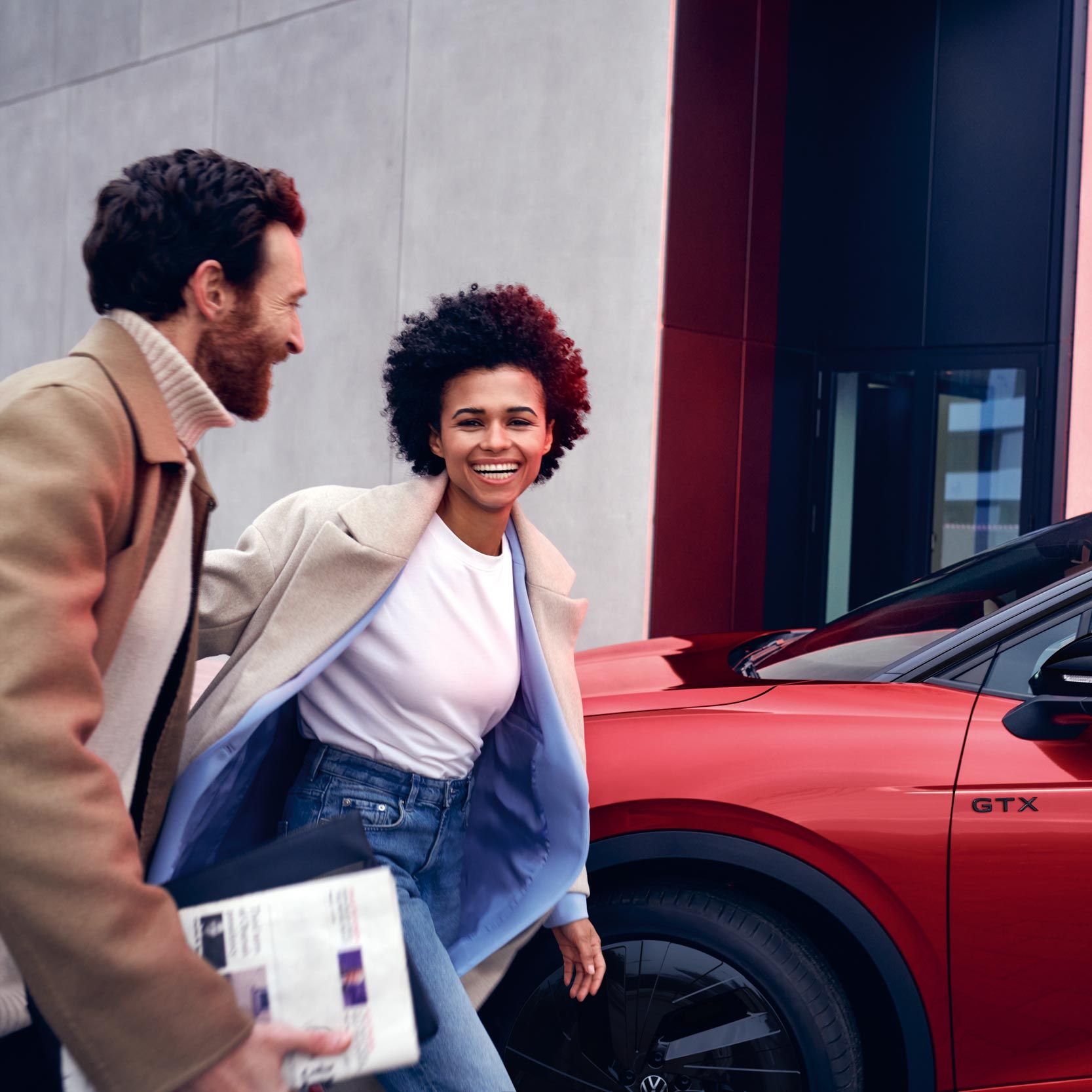 VW ID.4 GTX in Rot von der Seite am Straßenrand geparkt. Frau und Mann gehen lachend am Fahrzeug vorbei.