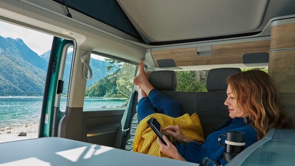 Eine Dame entspannt in einem VW California 6.1 und surft am Handy