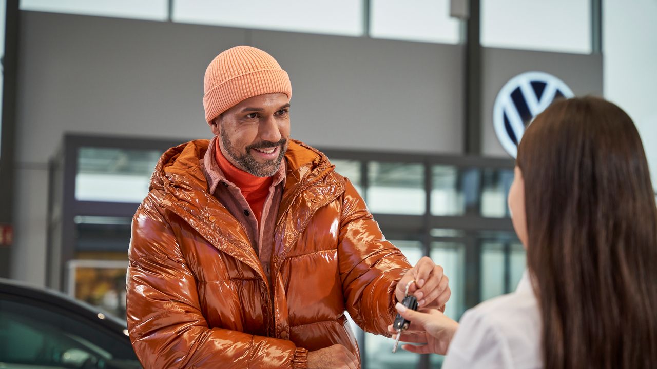Ein Mann in orangener Jacke gibt Autoschlüssel beim Händler ab