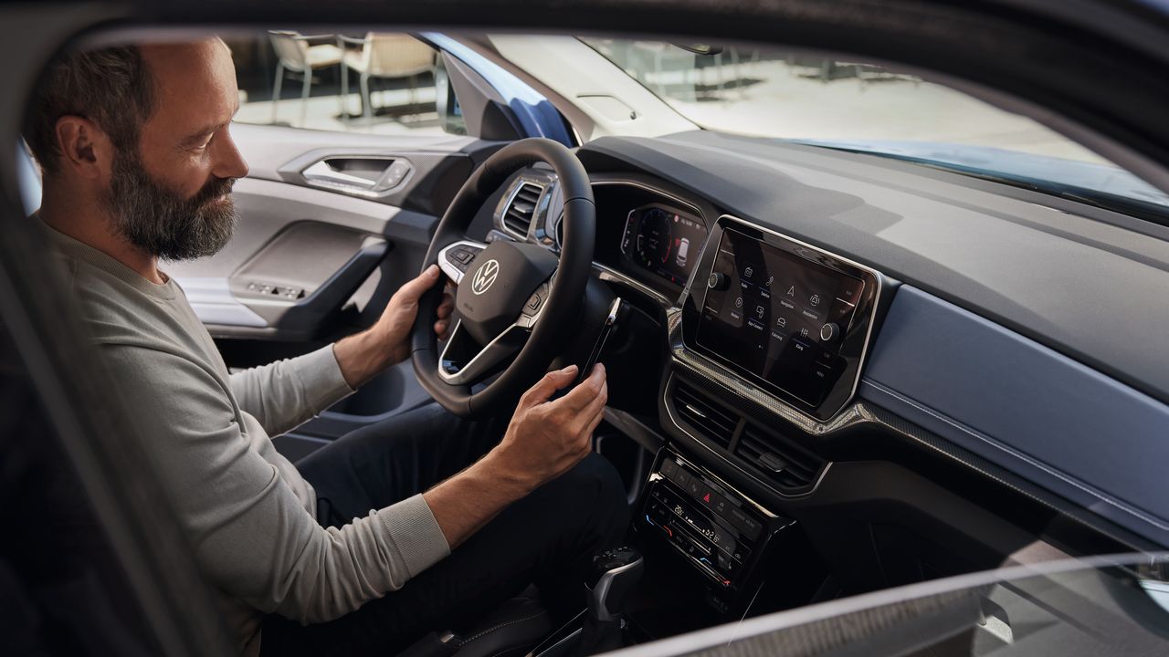 Ein Mann schaut auf dem Fahrersitz bei geöffneter Tür aufs Handy im VW T-Cross