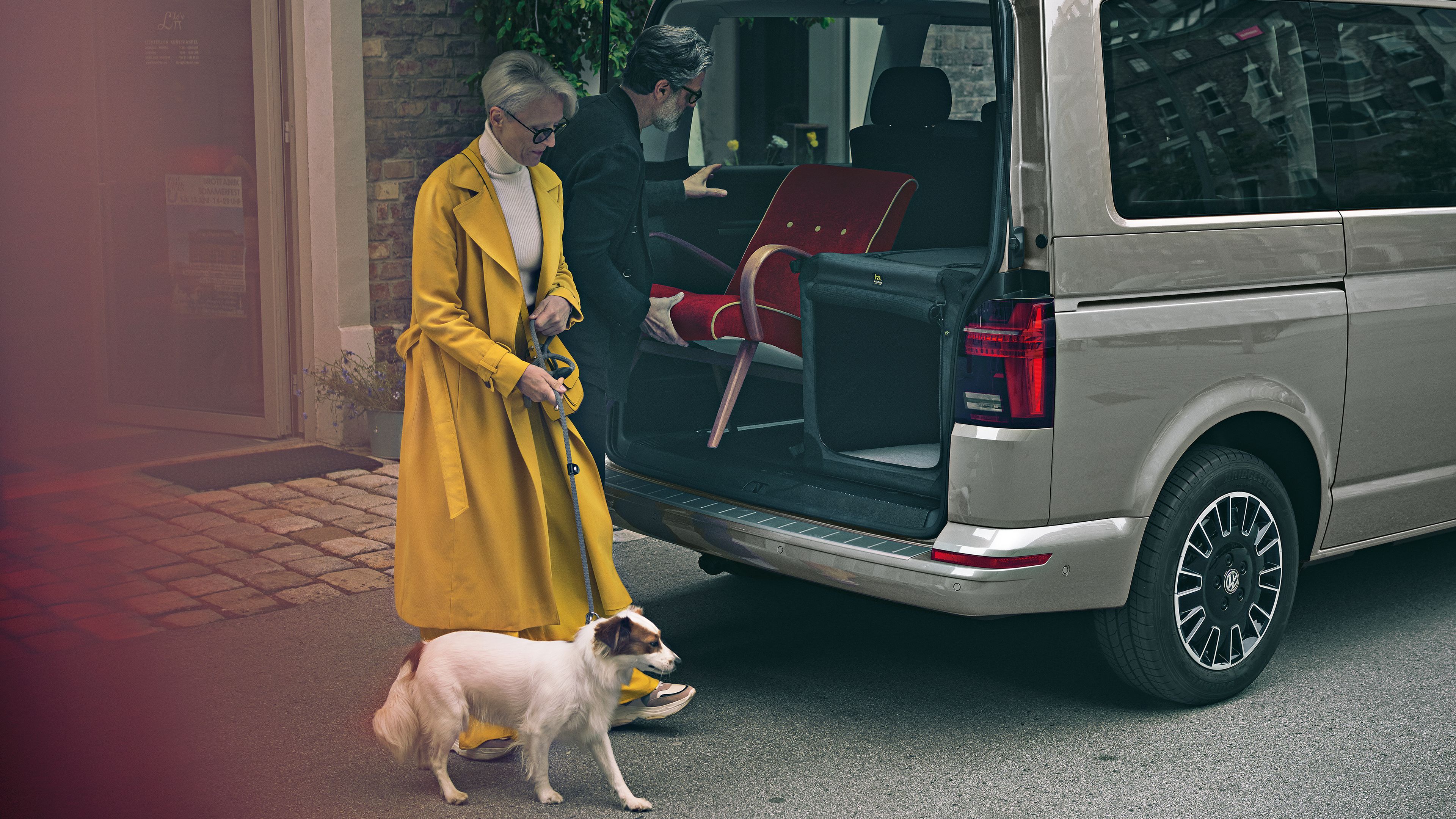 Ein Mann belädt den Kofferraum des VW Multivan 6.1, eine Frau mit Hund geht daran vorbei. 