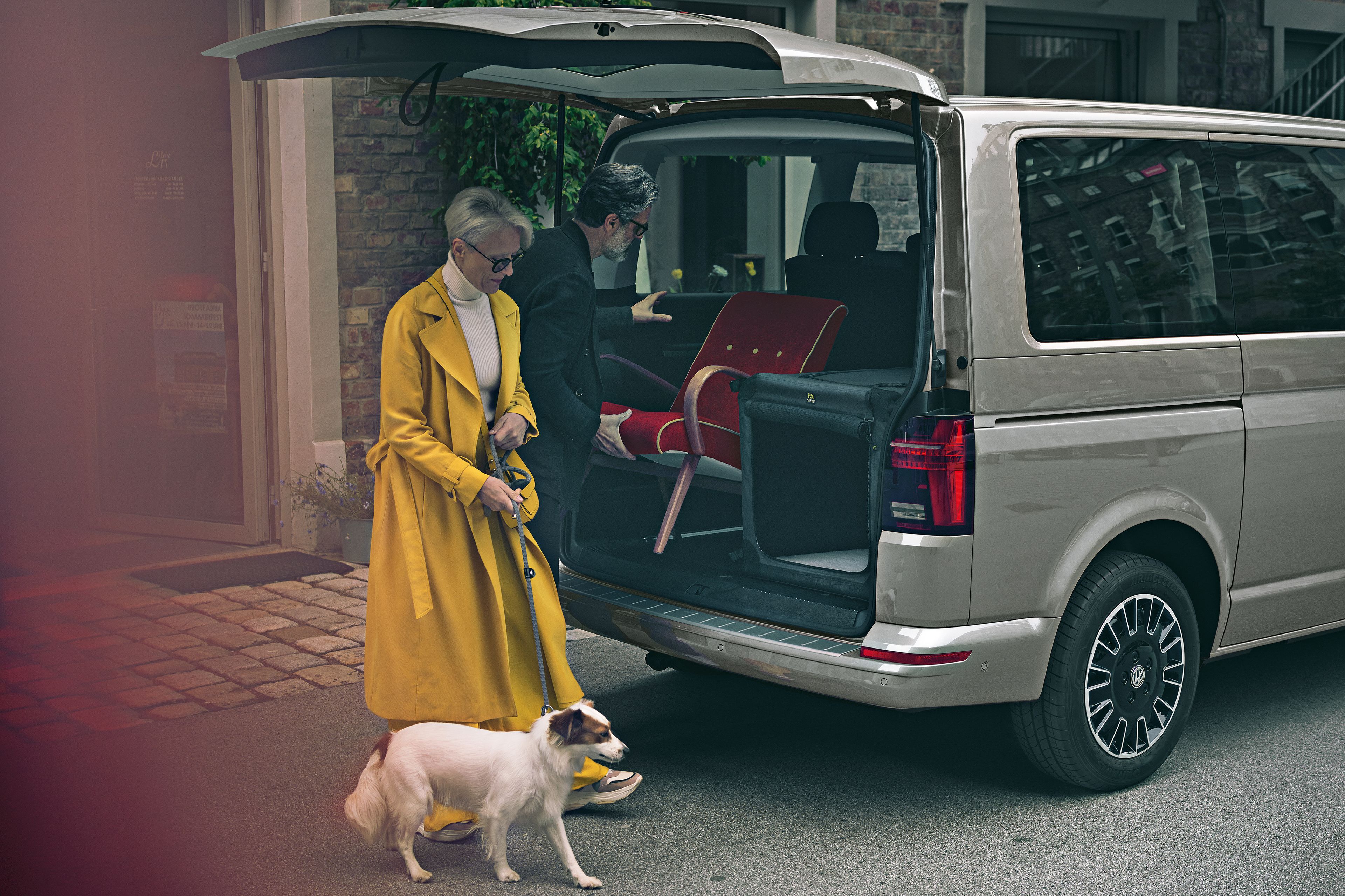 Ein Mann belädt den Kofferraum des VW Multivan 6.1, eine Frau mit Hund geht daran vorbei. 