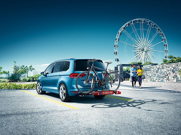 Ein VW Touran in blau mit Fahrradträger parkt auf einem Parkplatz, im Hintergrund steht ein Riesenrad.