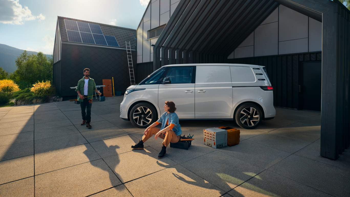 Ein weißer Volkswagen ID. Buzz Cargo vor einer Werkshalle, im Vordergrund sind ein Mann und eine Frau zu sehen