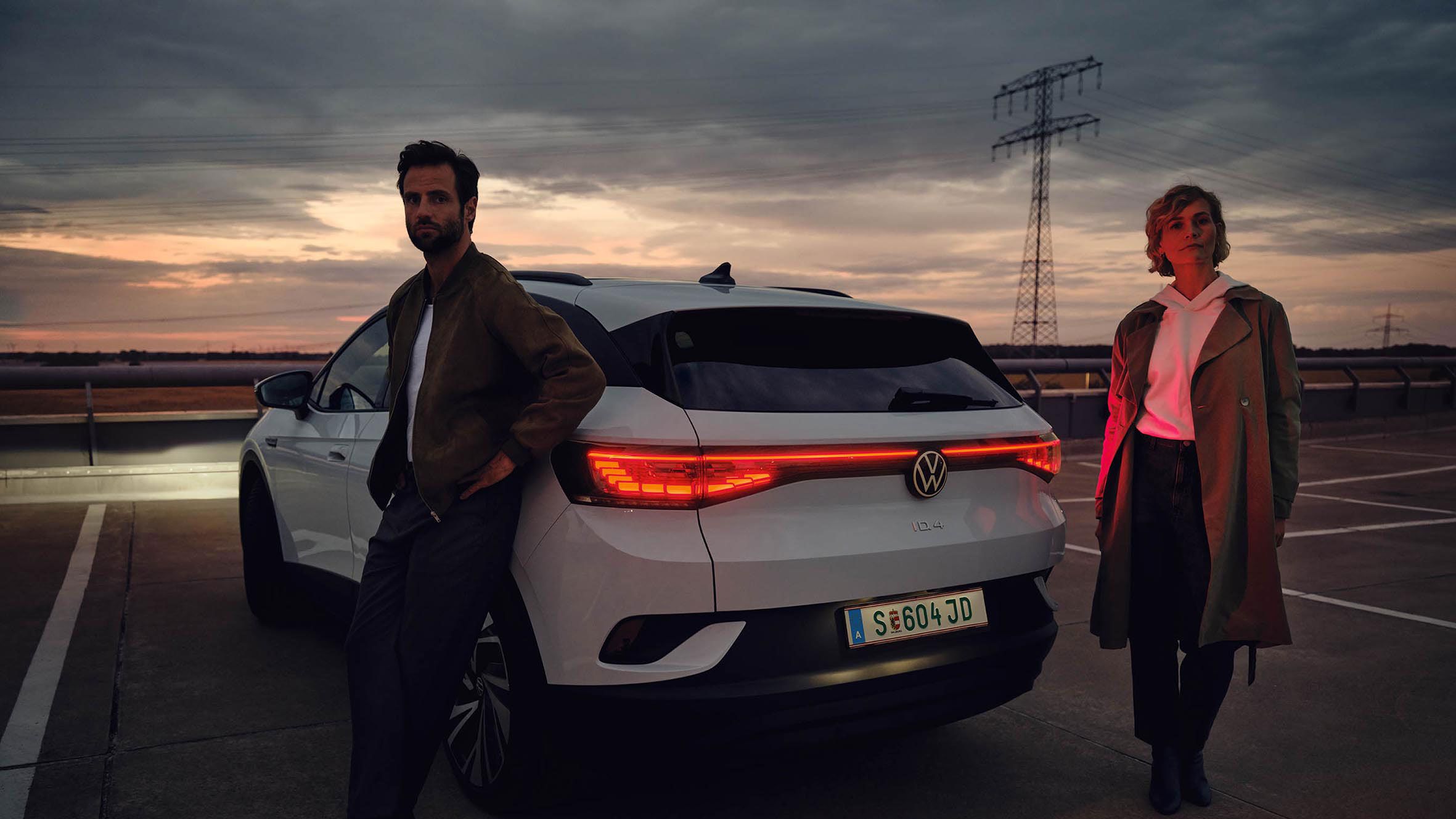 Frau und Mann stehen neben dem Heck eines weißen VW ID.4 bei Dämmerung auf einem Parkplatz