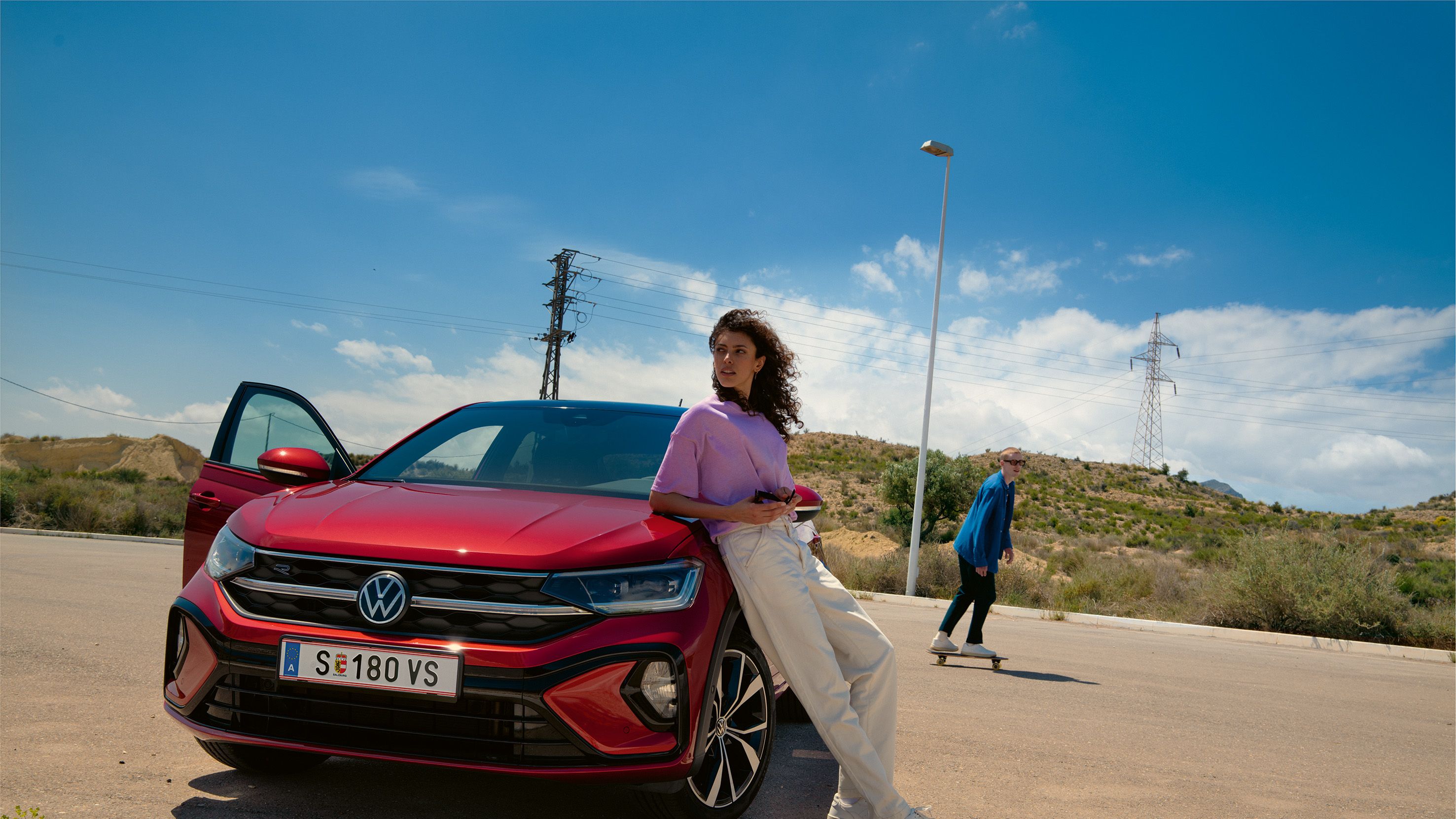 VW Taigo R-Line in Rot, Frontansicht auf Parkplatz. Frau lehnt an Motorhaube, Mann fährt Skateboard