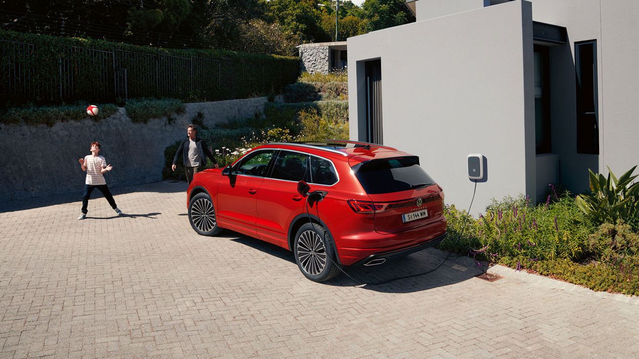 Ein VW Touareg Elegance eHybrid, Ansicht von schräg hinten, an einer Ladesäule vor einem modernen Haus.