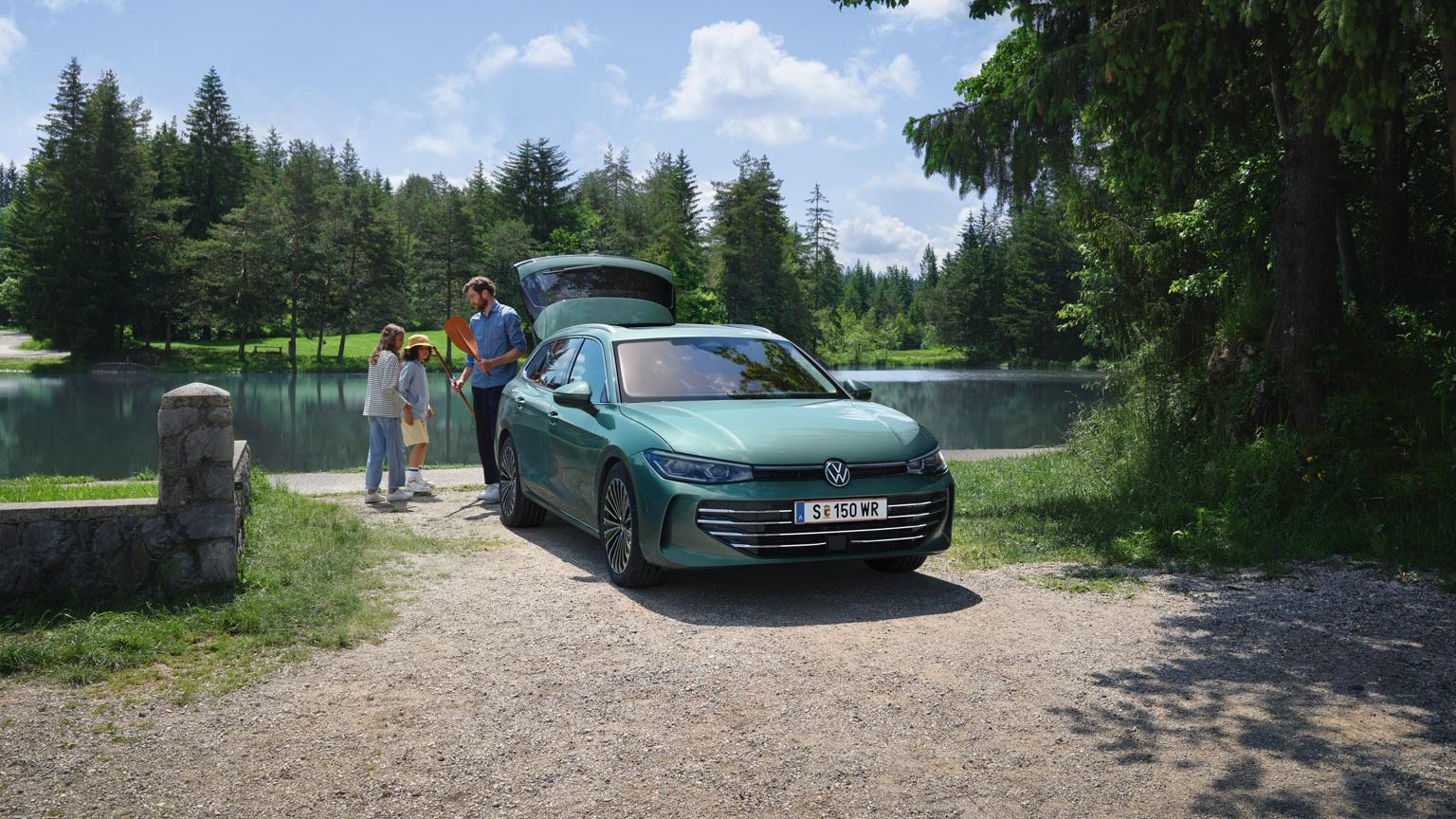 Der neue VW Passat in blaugrün parkt an einem Badesee mit geöffnetem Kofferraum. Ein Mann verteilt Freitzeitspielzeug an zwei Kinder.