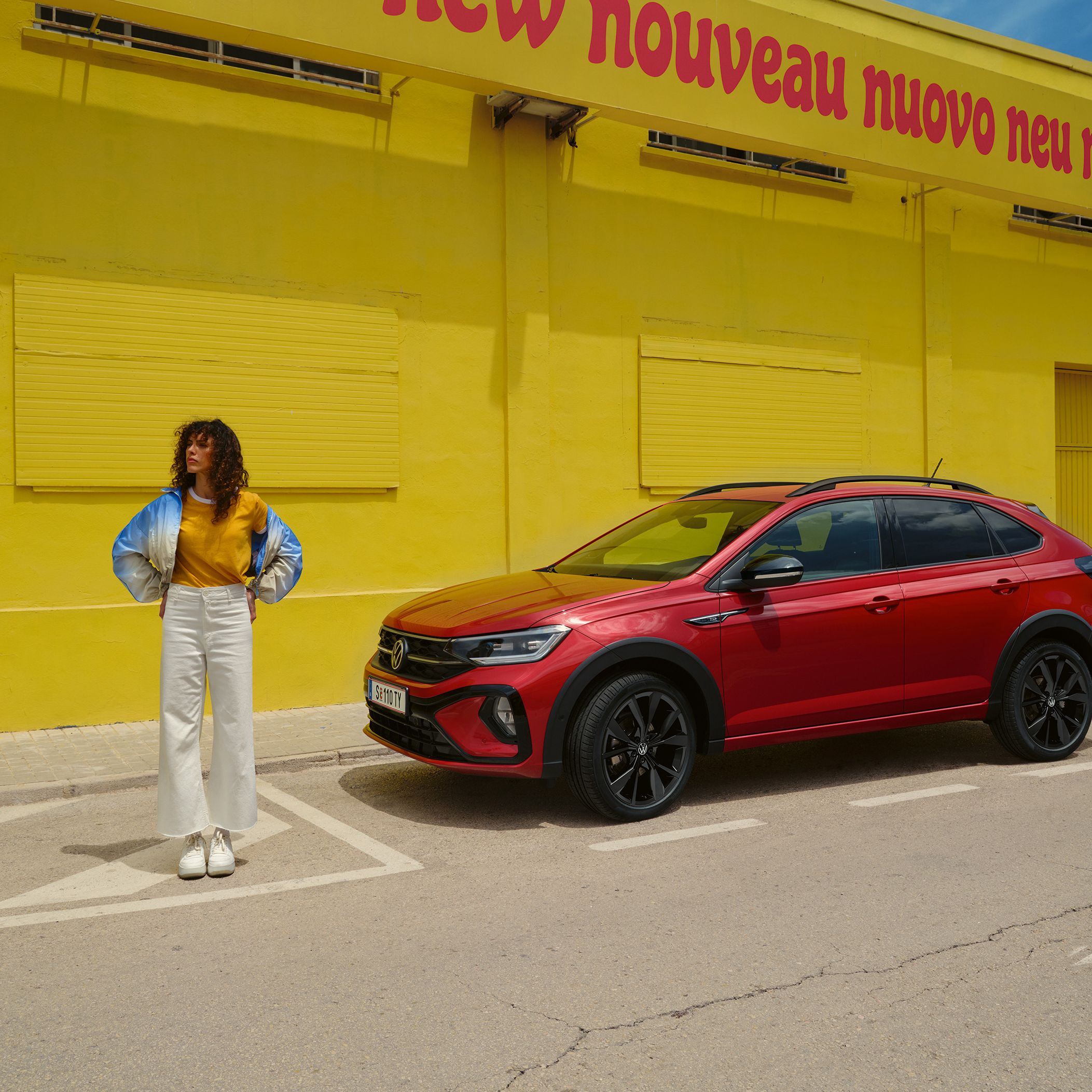 Der VW Taigo R-Line in Rot am Straßenrand vor gelbem Gebäude, Seitenansicht, Frau steht neben Fahrzeug
