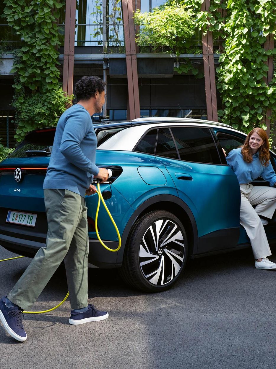 Ein Mann mit einem gelben Ladekabel für Elektroautos geht auf das Heck eines parkenden blauen VW ID.4 zu. Aus der Beifahrertür steigt eine junge Frau aus.