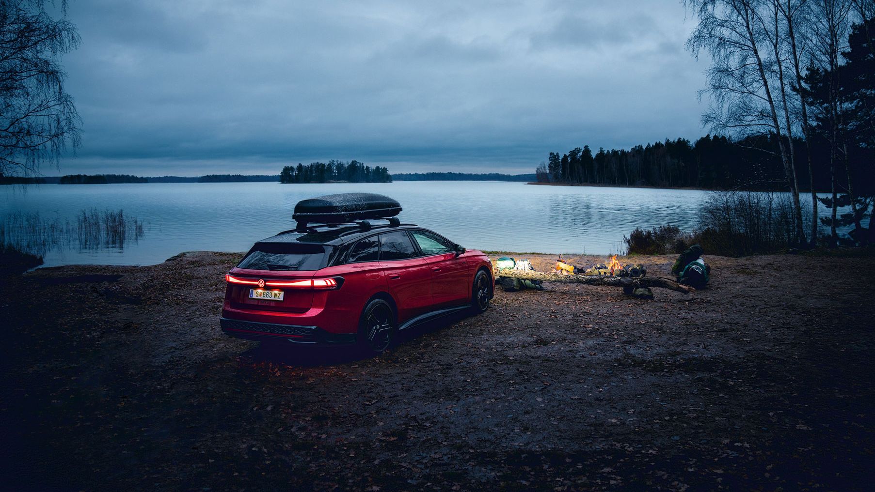Ein roter VW ID.7 Tourer GTX mit Dachbox vor einem See und Lagerfeuer