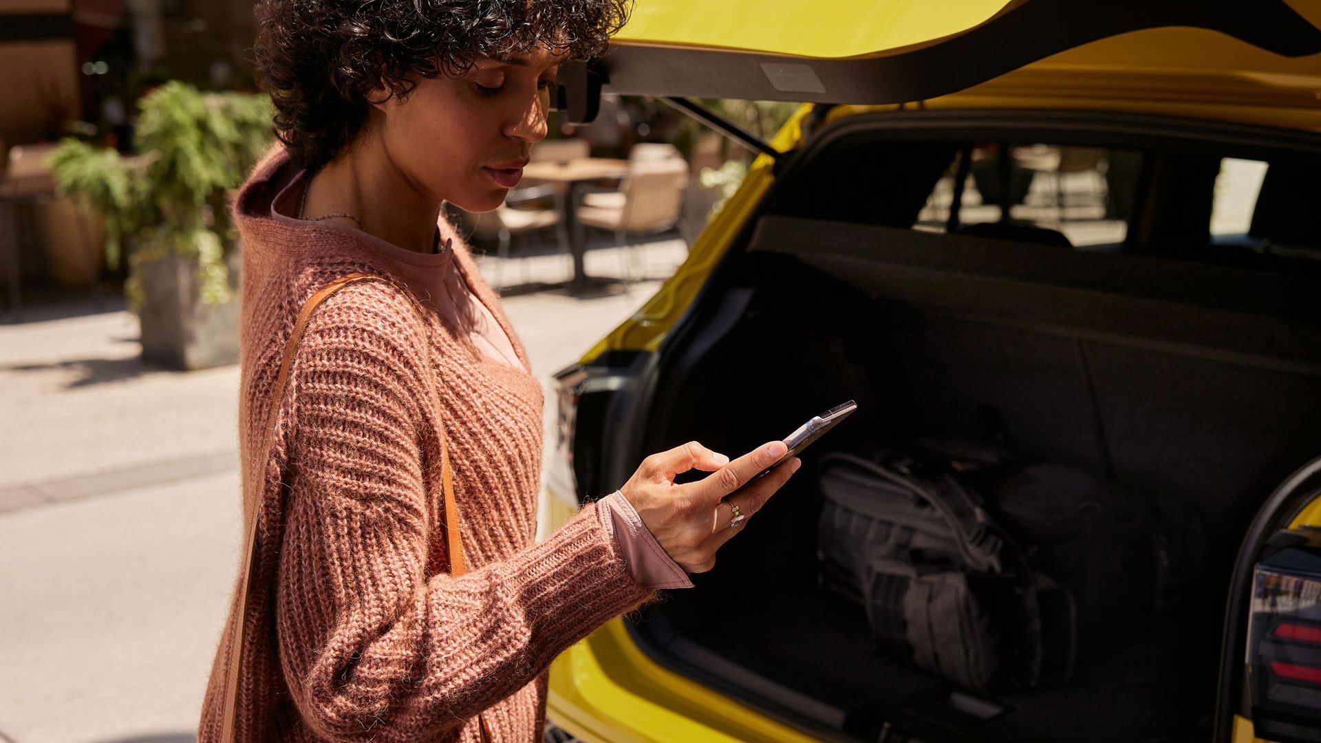 Eine frau schließt den Kofferraum eines gelben VW T-Cross und blickt dabei auf ihr Smartphone. 