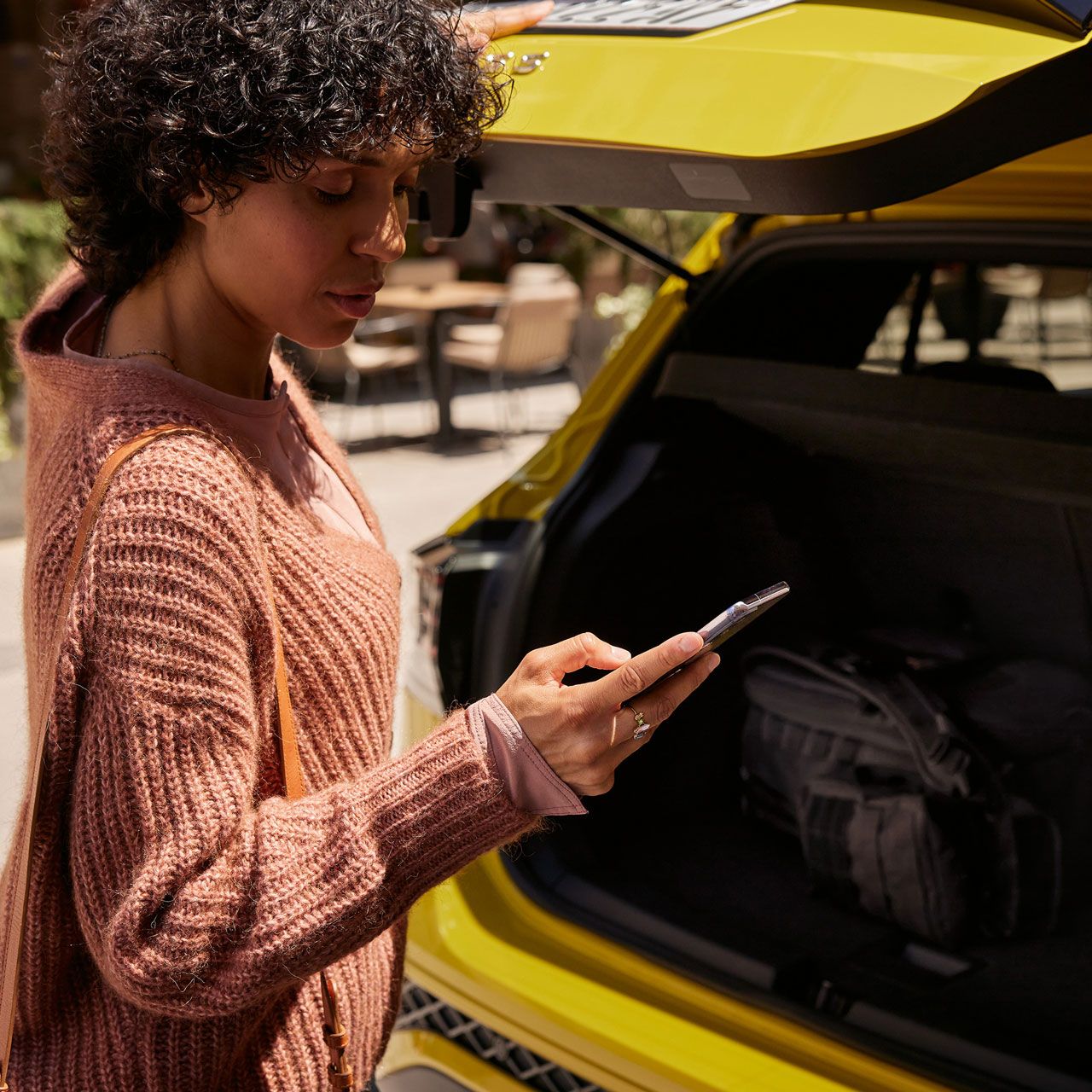Eine frau schließt den Kofferraum eines gelben VW T-Cross und blickt dabei auf ihr Smartphone. 