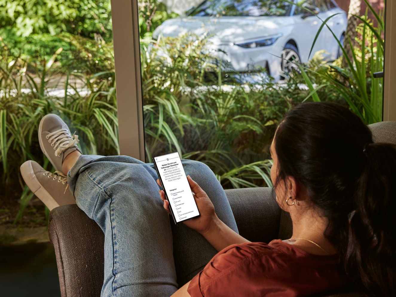 Eine Frau sitzt entspannt im Sofa und blickt auf ihr Smartphone, im Hintergrund ein VW ID.7