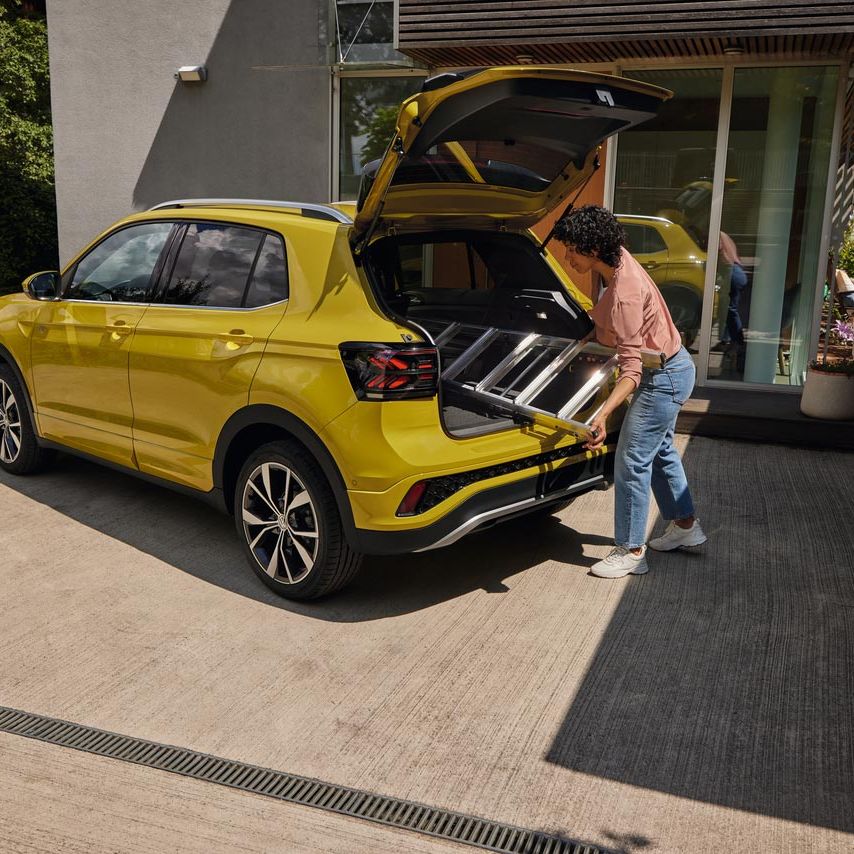 Eine junge Frau schiebt eine Leiter in den Kofferraum eines gelben VW T-Cross.