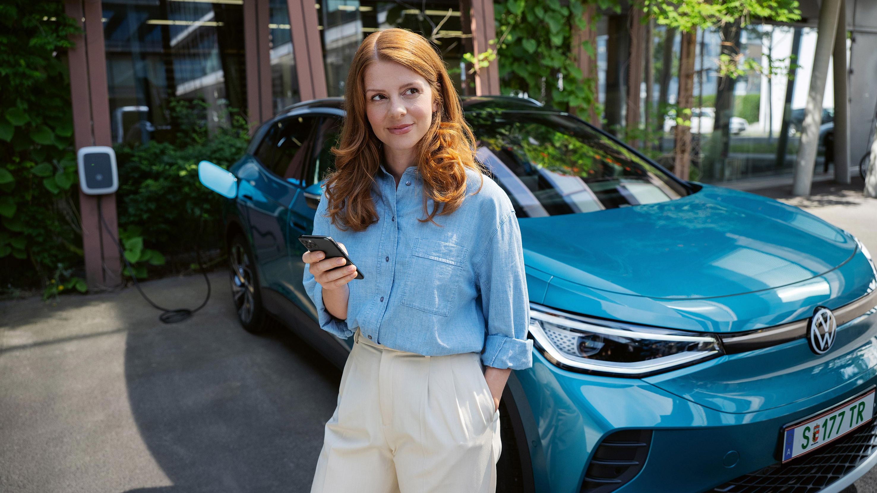 Ein blauer ID.4 wird aufgeladen, eine Frau steht vor dem Auto mit einem Smartphone in der Hand