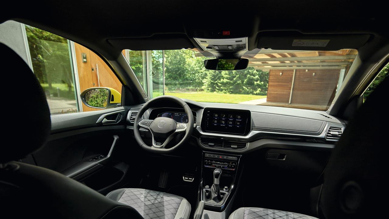 Blick vom Rücksitz aus ins Cockpit eines VW T-Cross R-Line und durch die Frontscheibe auf ein Haus mit Garten.