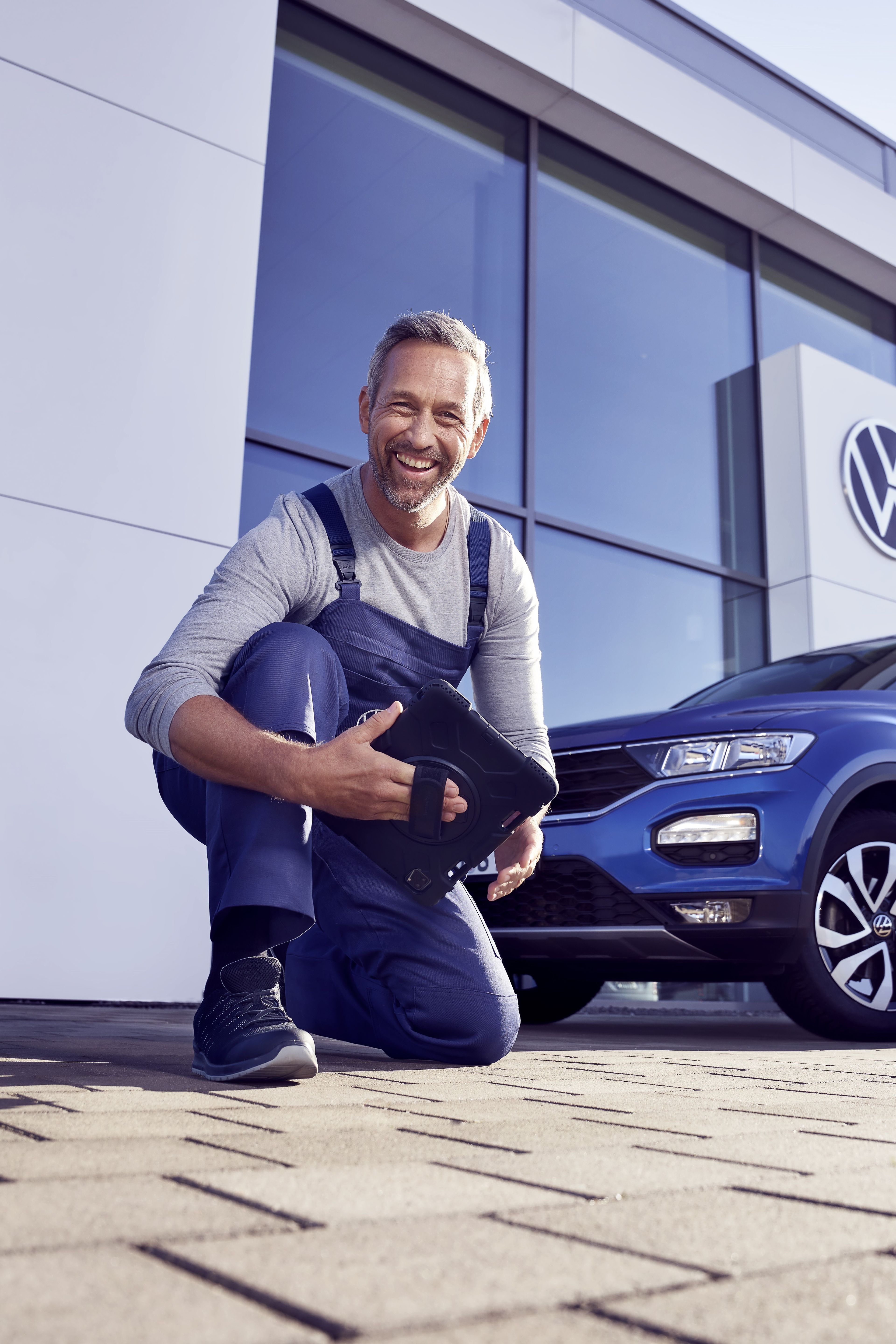 Ein Volkswagen Service Techniker mit Tablet, kniet vor einem blauen Volkswagen T-Roc