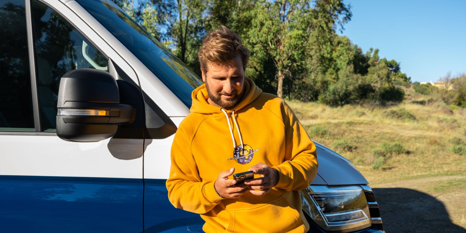 Ein Mann lehnt an seinem VW Nutzfahrzeug und blickt auf sein Smartphone