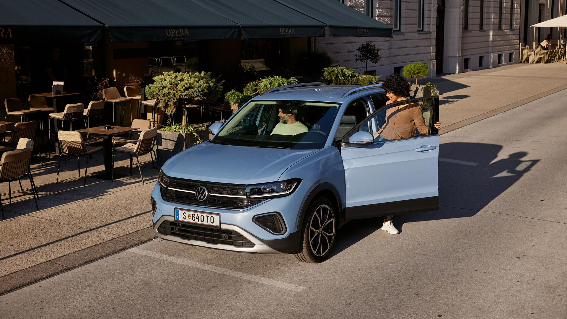Eine Frau steigt auf der Fahrerseite in den blauen VW T-Cross Style, der vor einem urbanen Straßencafé parkt.