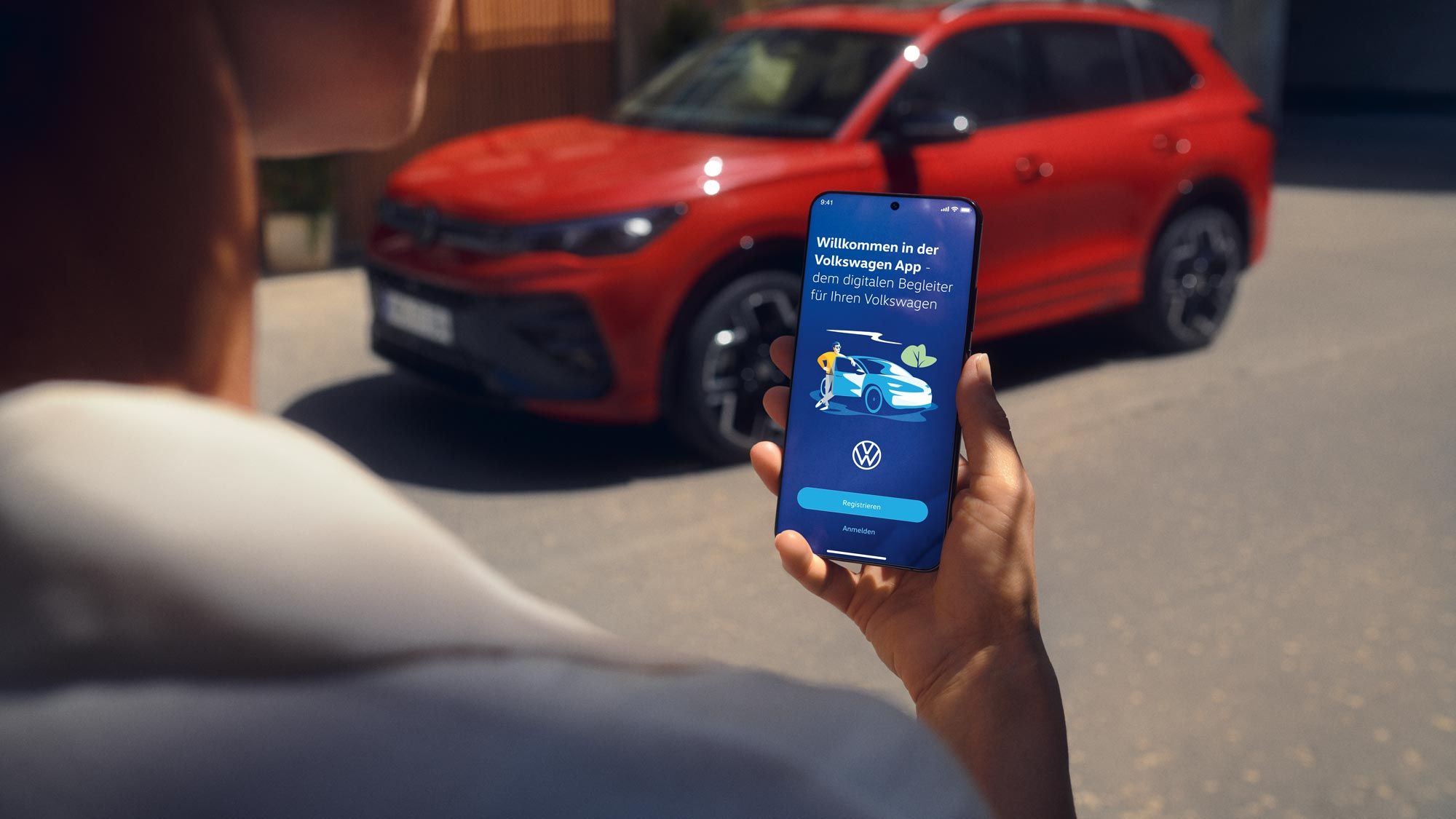 Blick auf ein Handydisplay mit Fahrzeugdaten des Tiguan, im Hintergrund ein seitlich geparkter VW Tiguan in rot