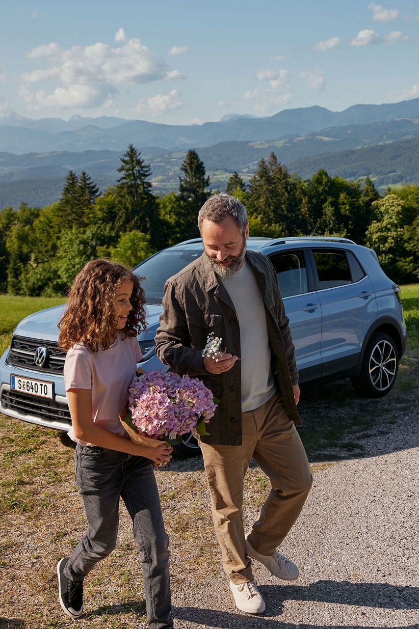 Ein blauer VW T-Cross steht geparkt auf einer Wiese in den Bergen. Davor laufen ein Mädchen und ein Mann mit Blumen in der Hand.