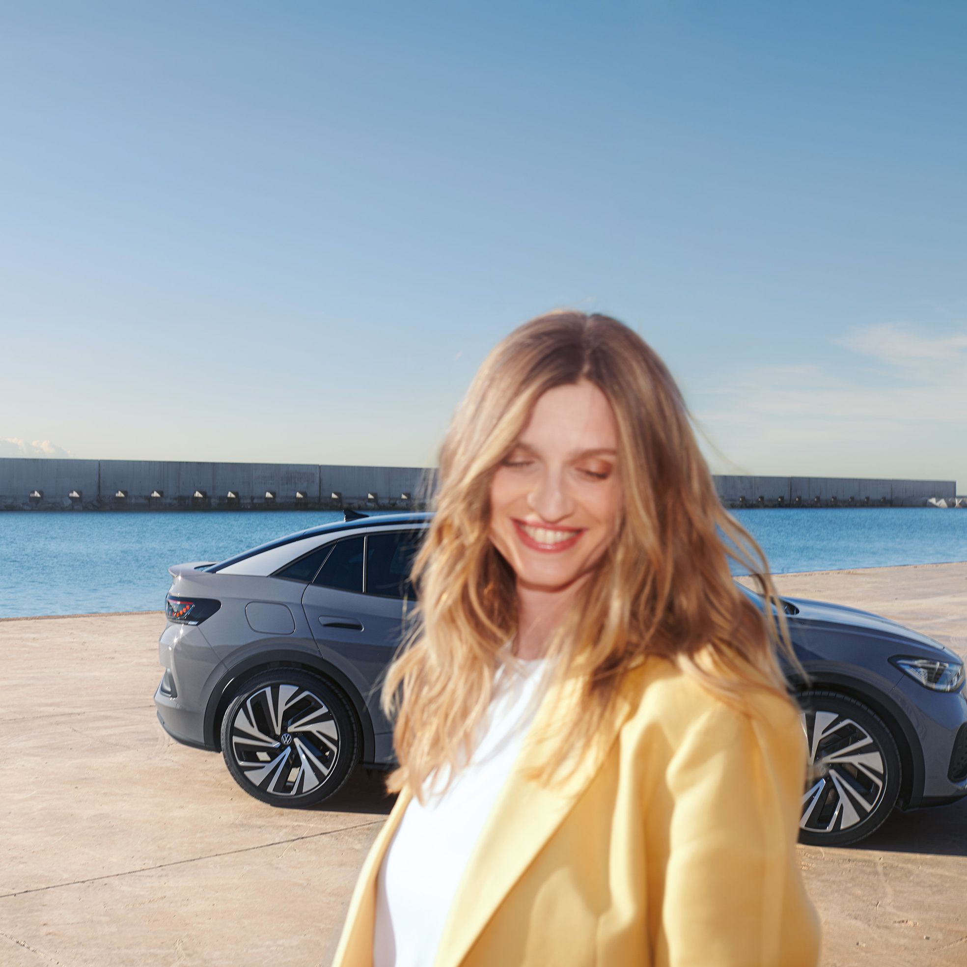 Seitenansicht eines grauen VW ID.5 an einem Hafenkai, im Vordergrund eine lächelnde, hell gekleidete Frau