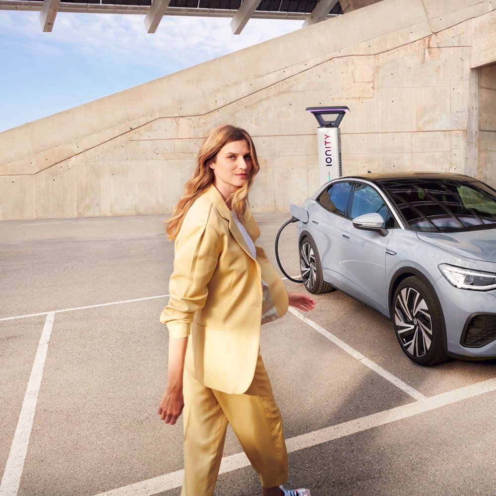 VW ID.5 in Grau steht auf einem Parkplatz und lädt an einer Ladesäule, Sicht auf Front und Seite, Frau geht auf Fahrzeug zu