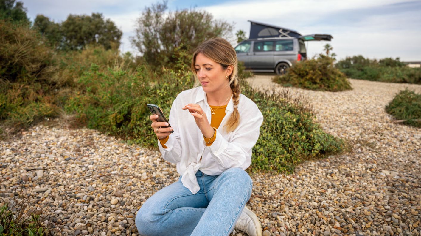 Eine Frau sitzt im Freien am Boden und blickt auf ihr Smartphone, im Hintergrund ein VW California