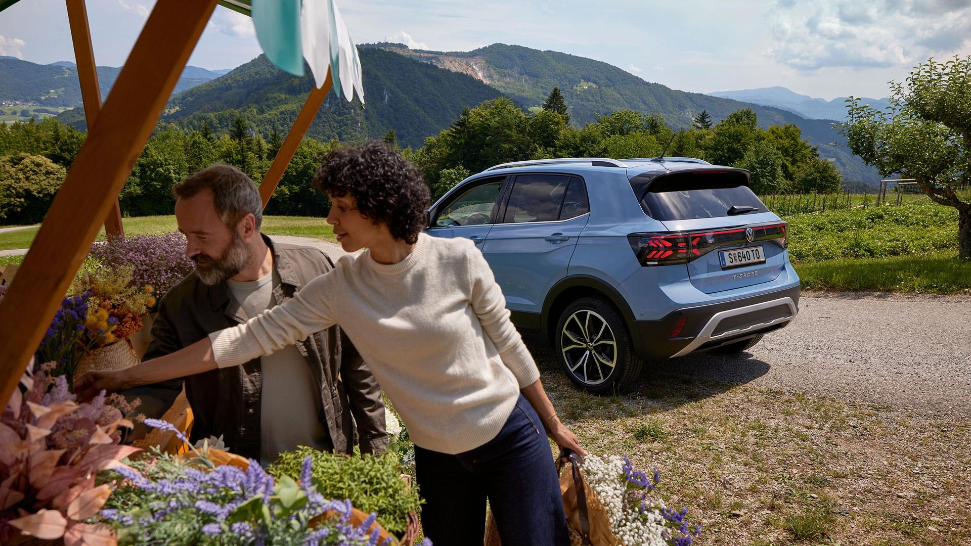 Mann und Frau stehen bei einem Blumenstand, hinter ihnen ein blauer VW T-Cross vor bergiger Landschaft.