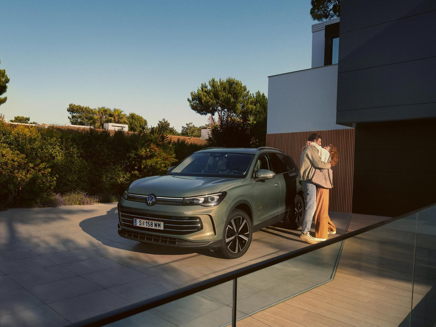 Eine Frau und ein Mann umarmen sich, neben ihnen steht ein VW Tiguan Elegance in der Farbe Cipressino-Grün Metallic