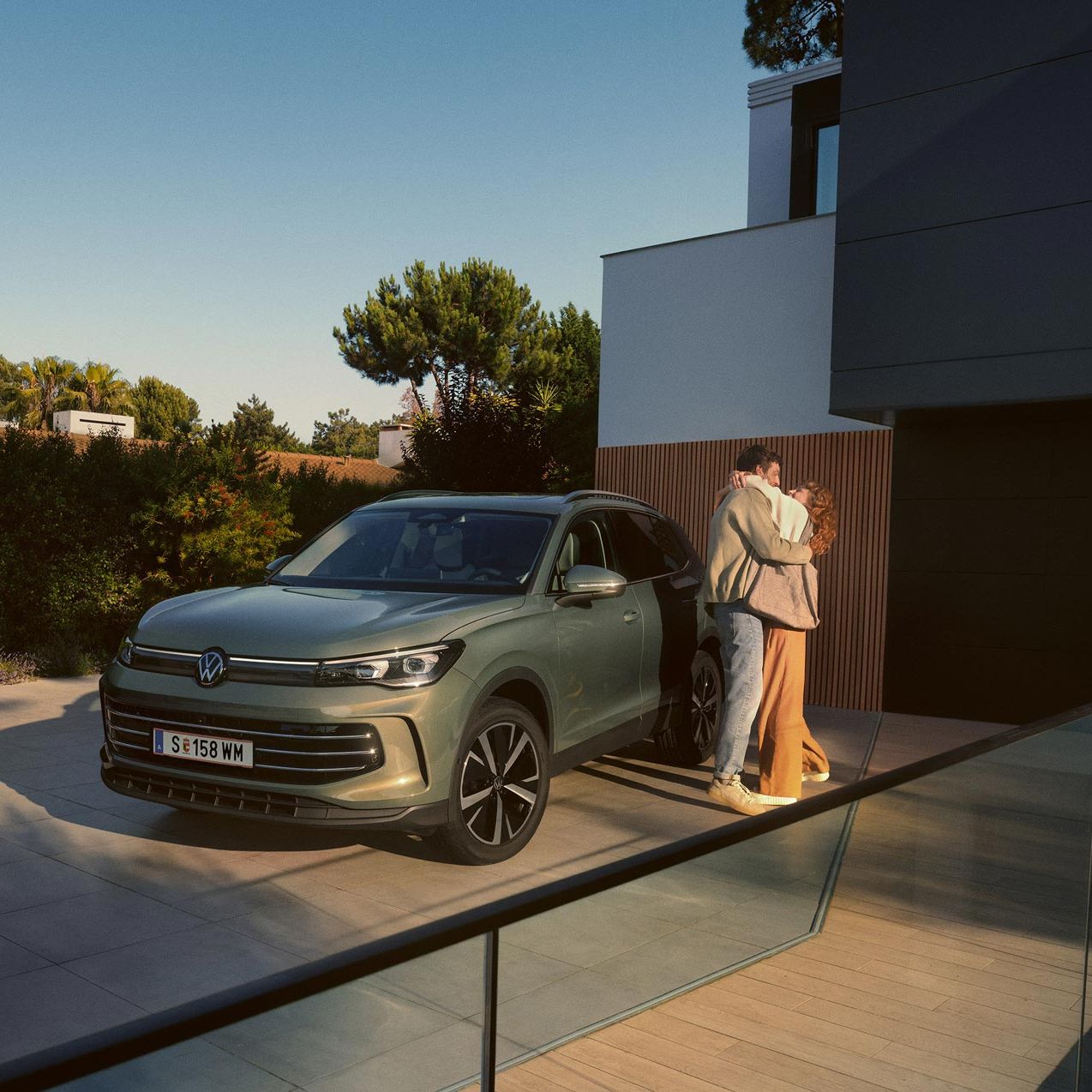 Eine Frau und ein Mann umarmen sich, neben ihnen steht ein VW Tiguan Elegance in der Farbe Cipressino-Grün Metallic