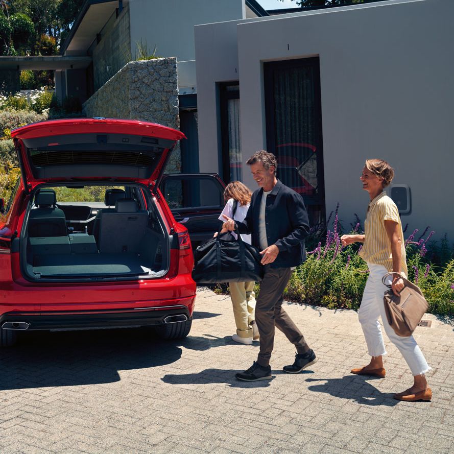 Ein Mann und eine Frau gehen auf einen VW Touareg Elegance eHybrid mit geöffnetem Kofferraum zu.