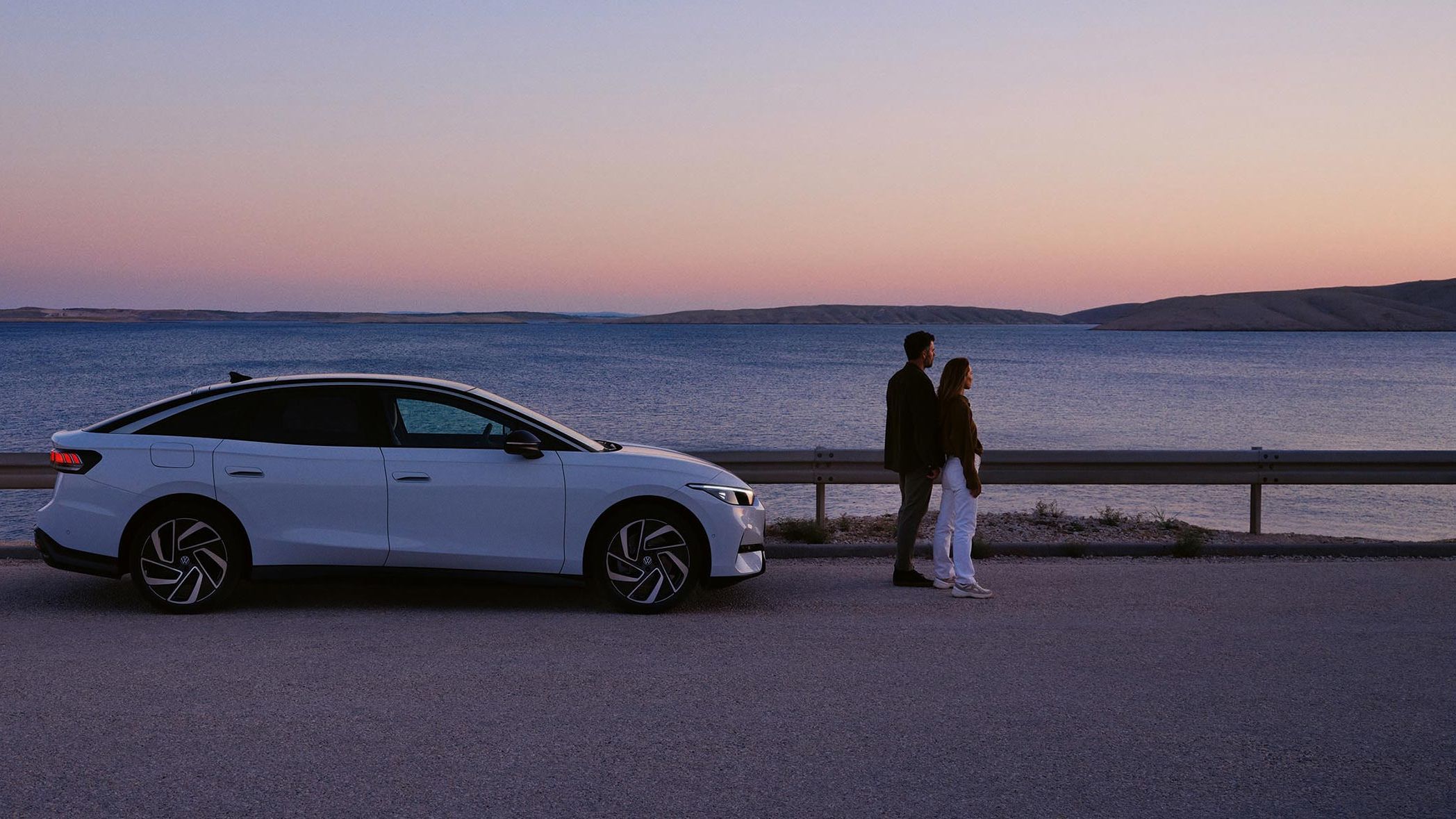 Ein VW ID.7 steht geparkt am Meer. Vor dem Auto steht ein Pärchen, das in den Sonnenuntergang blickt.