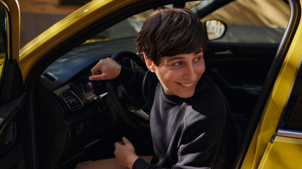 Eine junge Frau schaut auf dem Fahrersitz eines gelben VW T-Cross durch die geöffnete Tür lächelnd über die Schulter.
