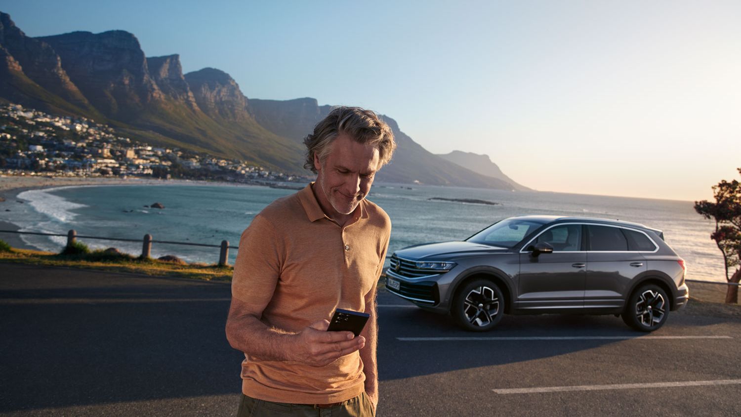 Ein Mann schaut auf sein Smartphone, dahinter parkt ein VW Touareg Elegance, im Hintergrund das Meer und die Küste.