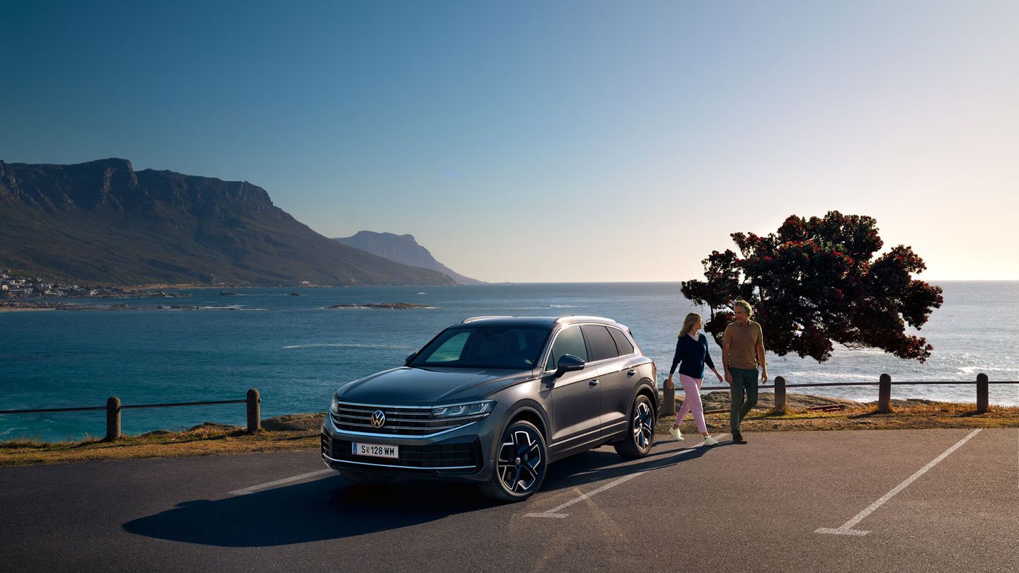 Der neue Volkswagen Touareg auf einem Parkplatz an der Küste, an ihm gehen eine Frau und ein Mann vorbei