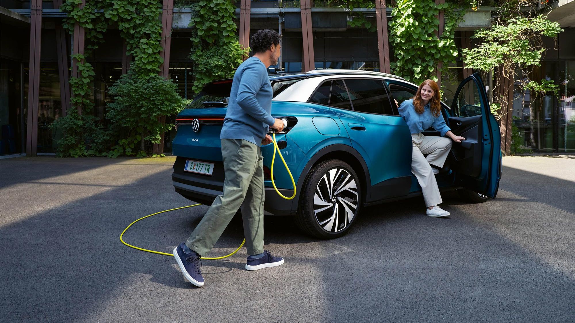 Ein Mann mit einem gelben Ladekabel für Elektroautos geht auf das Heck eines parkenden blauen VW ID.4 zu. Aus der Beifahrertür steigt eine junge Frau aus.