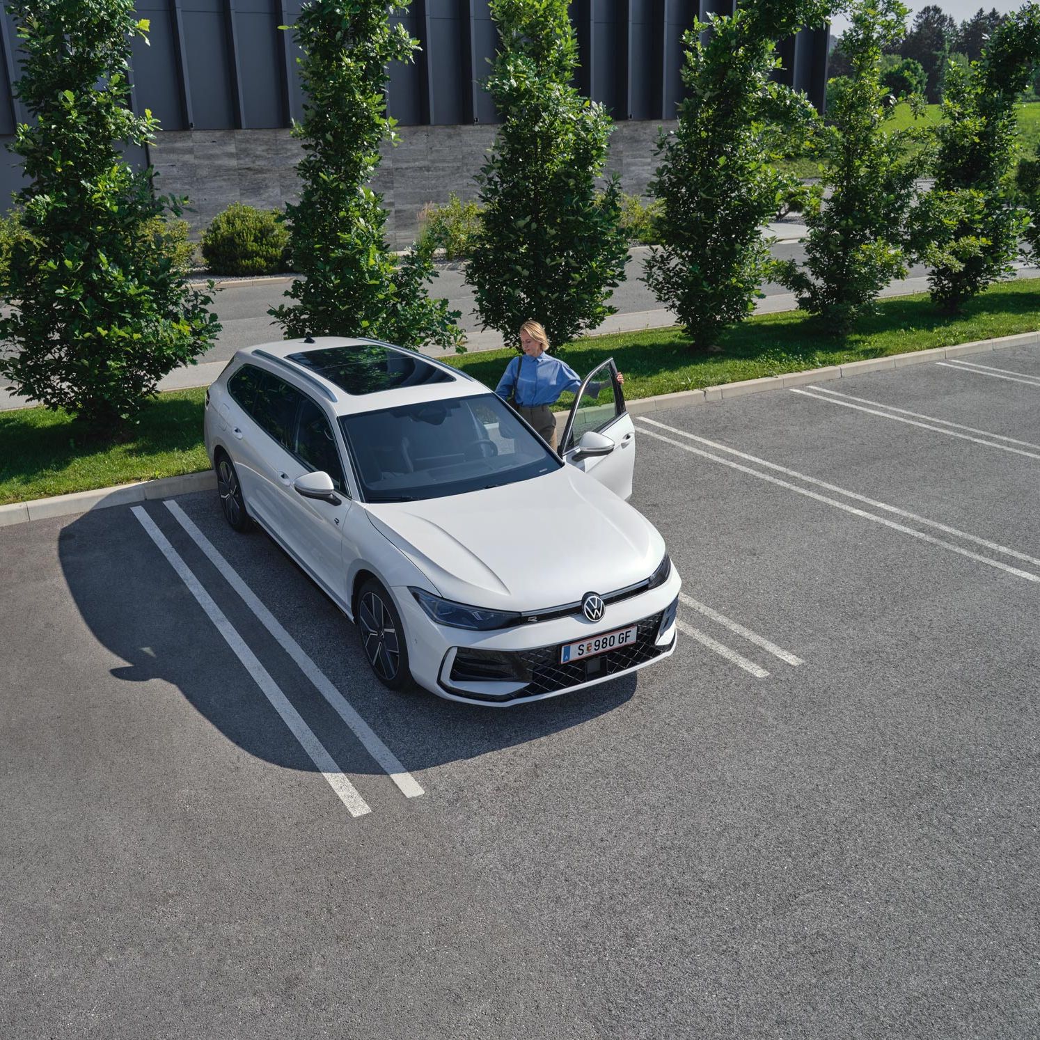 Topshot eines neuen weißen VW Passat auf einem Parkplatz, eine Frau steigt auf der Fahrerseite ein.