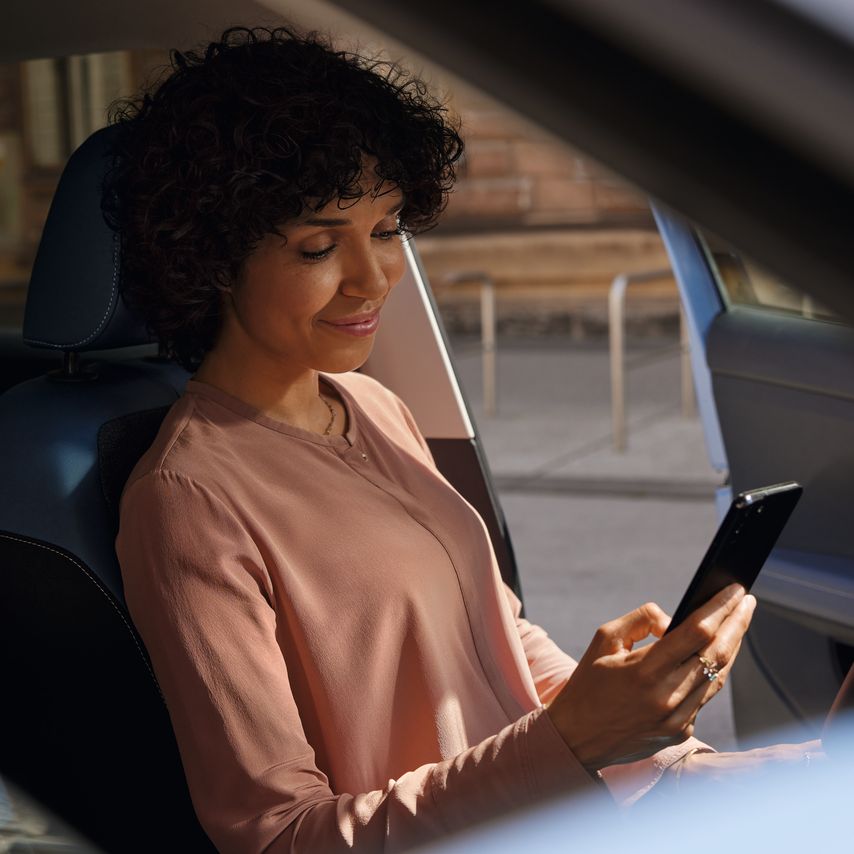 Eine lächelnde Frau schaut auf dem Fahrersitz eines geparkten VW T-Cross aufs Handy.