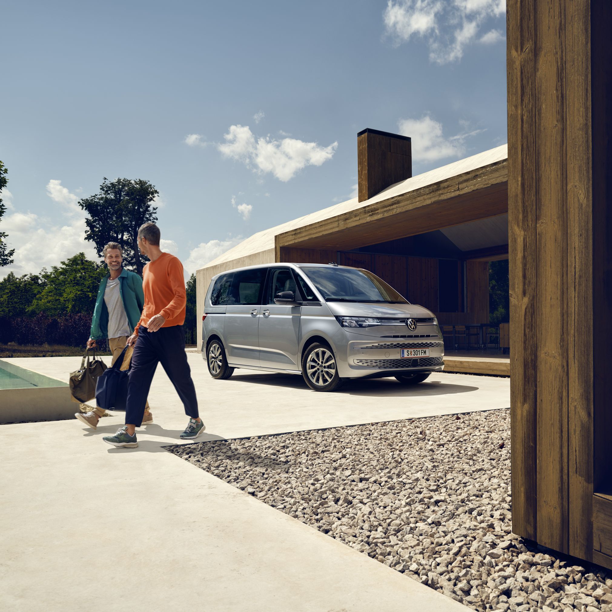 Ein Volkswagen Multivan in Silber parkt vor einem Haus mit Pool, zwei Männer mit Sporttaschen bewegen sich von dem Fahrzeug fort.