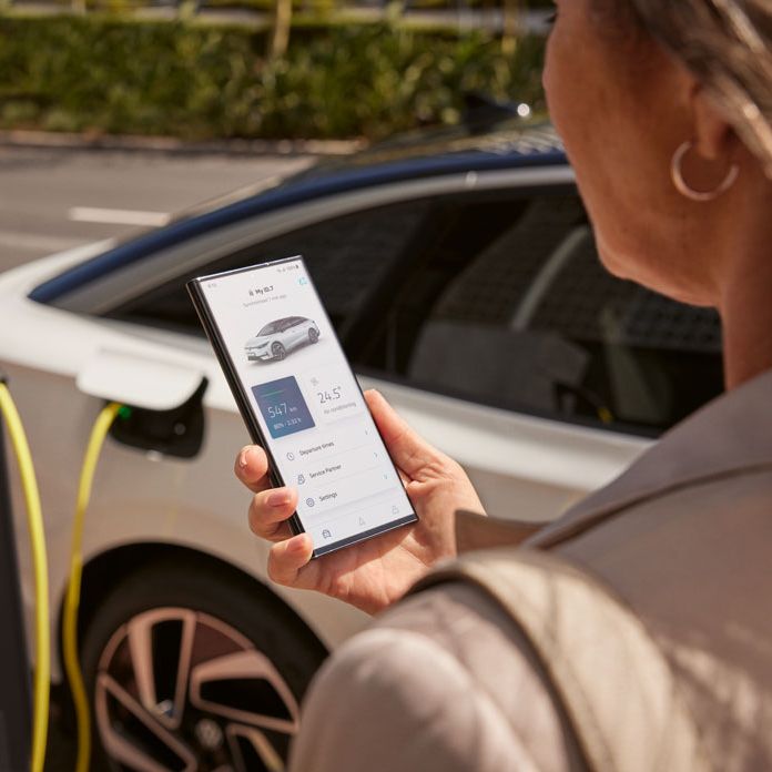 Ein VW ID.7 beim Ladevorgang mittels Plug and Charge, Frau blickt auf die VW Connect App auf ihrem Smartphone