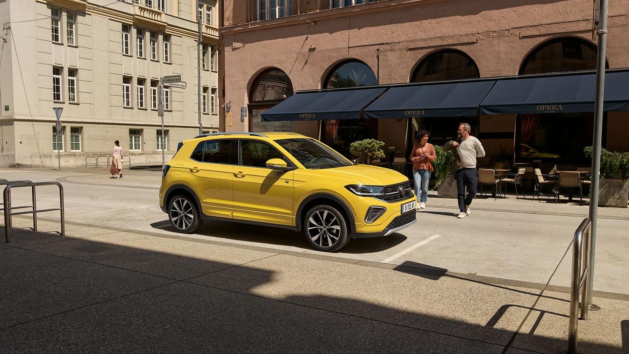 VW T-Cross in gelb parkt seitlich vor einem urbanen Straßencafé, ein Päarchen geht auf den Wagen zu.