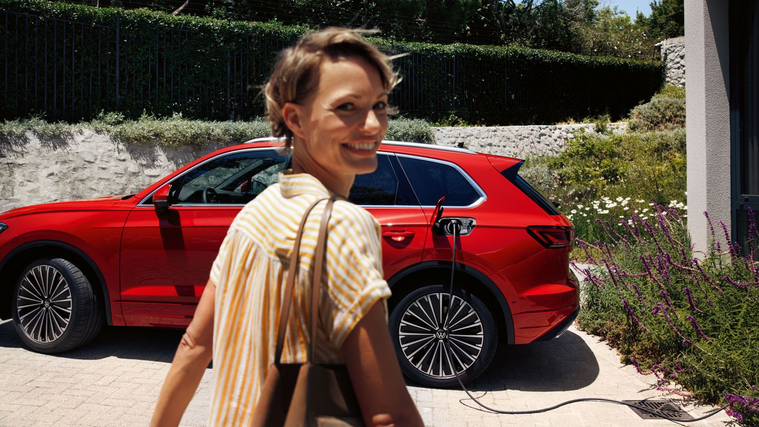 Im Vordergrund eine lächelnde Frau, im Hintergrund lädt ein VW Touareg Elegance eHybrid an einer Ladesäule.