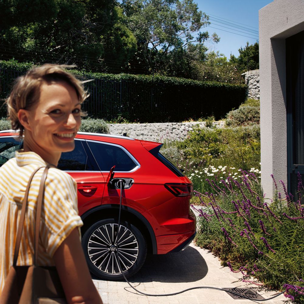 Im Vordergrund eine lächelnde Frau, im Hintergrund lädt ein VW Touareg Elegance eHybrid an einer Ladesäule.